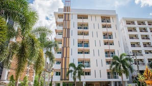 A multi-story hotel with a modern design, featuring a central vertical strip, balconies on each floor, and a parking area with a yellow car. - Napas Residence (Hat Yai)