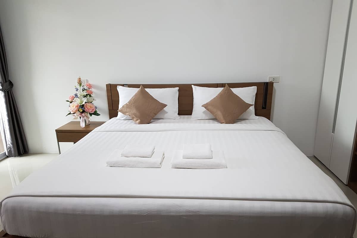 Una cama bien hecha con sábanas blancas y dos almohadas marrones, una mesita de noche con un florero, y una ventana con cortinas.