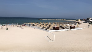 A beach with rows of sunbeds and umbrellas, a lifeguard tower, and a rocky breakwater in the distance. - Hotel New Belvedere (Mangalia)