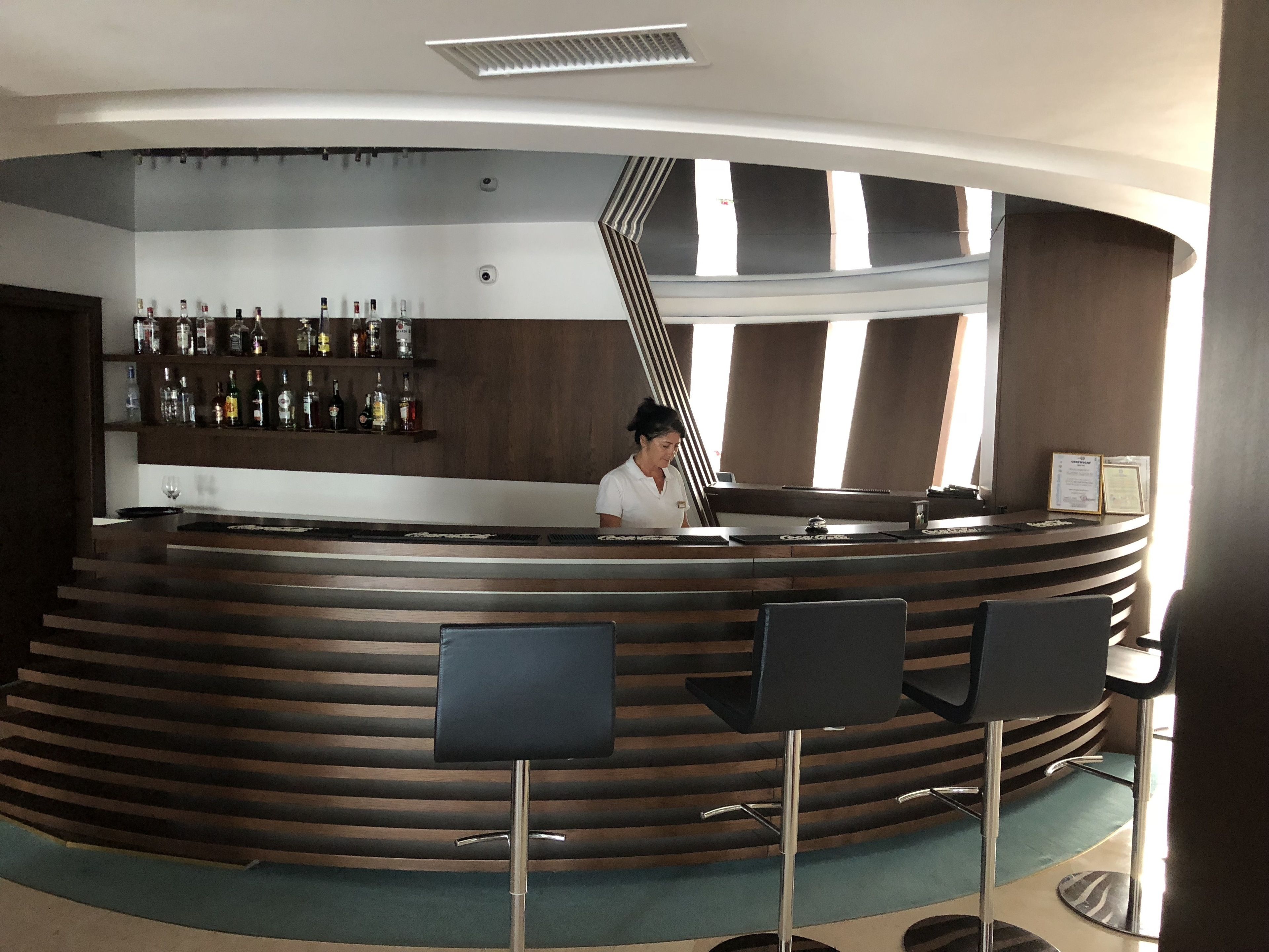 Ein gebogener Empfangstresen mit Barkeeper, eine Bar mit verschiedenen Spirituosenflaschen und eine Treppe, die zu einer oberen Ebene führt.