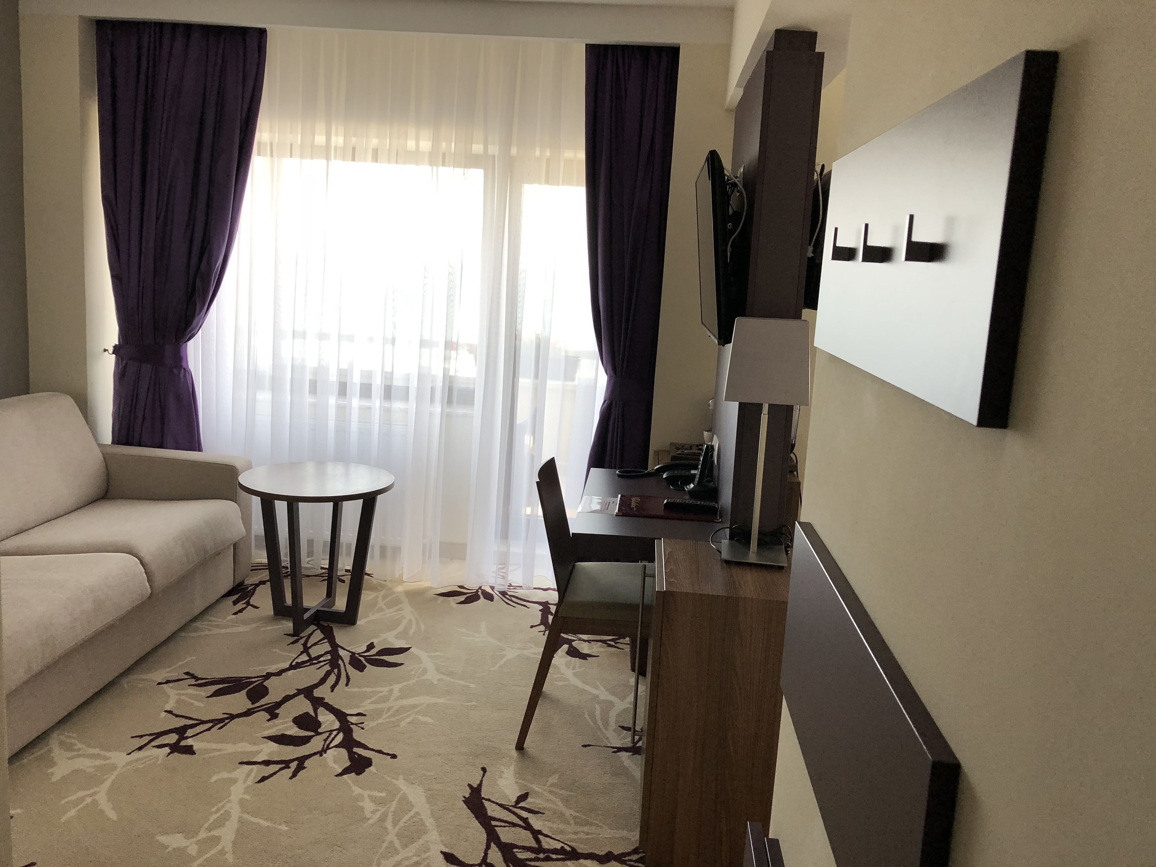 Habitación de hotel con sofá, una mesita redonda pequeña, escritorio con lámpara y vistas a través de cortinas translúcidas.