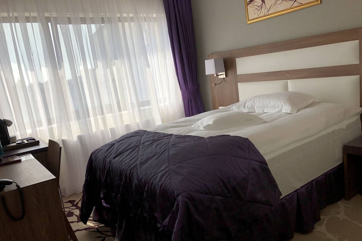 Habitació d'hotel amb llit, escriptori, cadira, tauleta de nit i finestra amb cortines transparents.