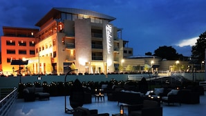 An outdoor seating area with multiple couches and tables, a large building with illuminated signage in the background, and a staircase leading to an upper level. - Hotel New Belvedere (Mangalia)