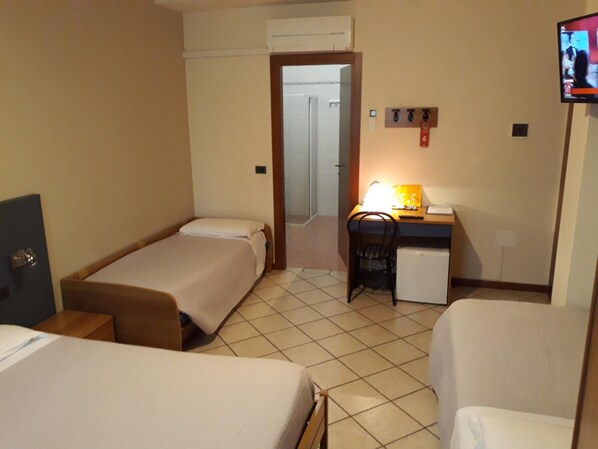 Habitación de hotel con dos camas individuales, un escritorio con silla, un refrigerador pequeño y un televisor en la pared.