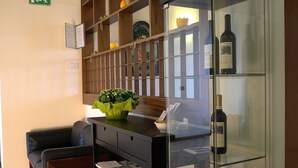 A reception area with a sofa, a desk with a vase of flowers, a glass cabinet with wine bottles, and a wooden shelving unit with decorative items.