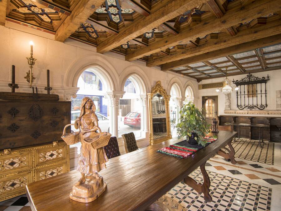 A dining area with a wooden table, chairs, and a statue. There are arched windows and a tiled floor.