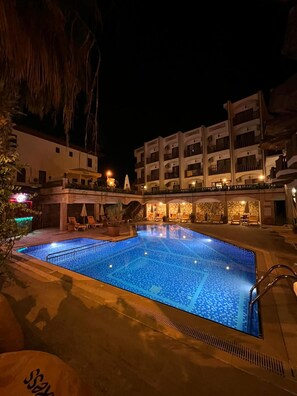 A swimming pool with a tiled pattern, surrounded by lounge chairs and a building with multiple balconies.