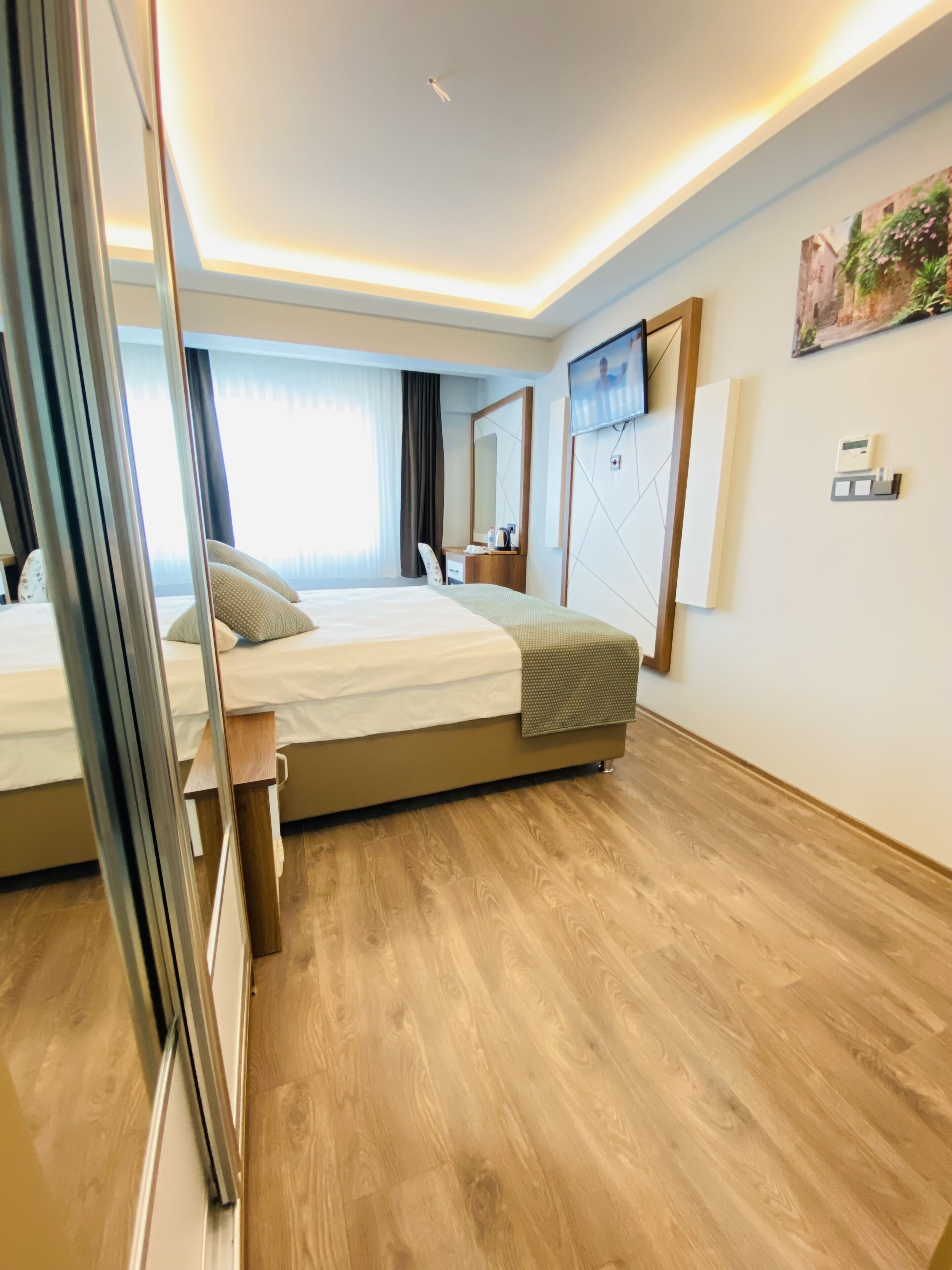 A modern hotel room with a large bed, wooden flooring, a mirror, a flat-screen TV, and a small bedside table.