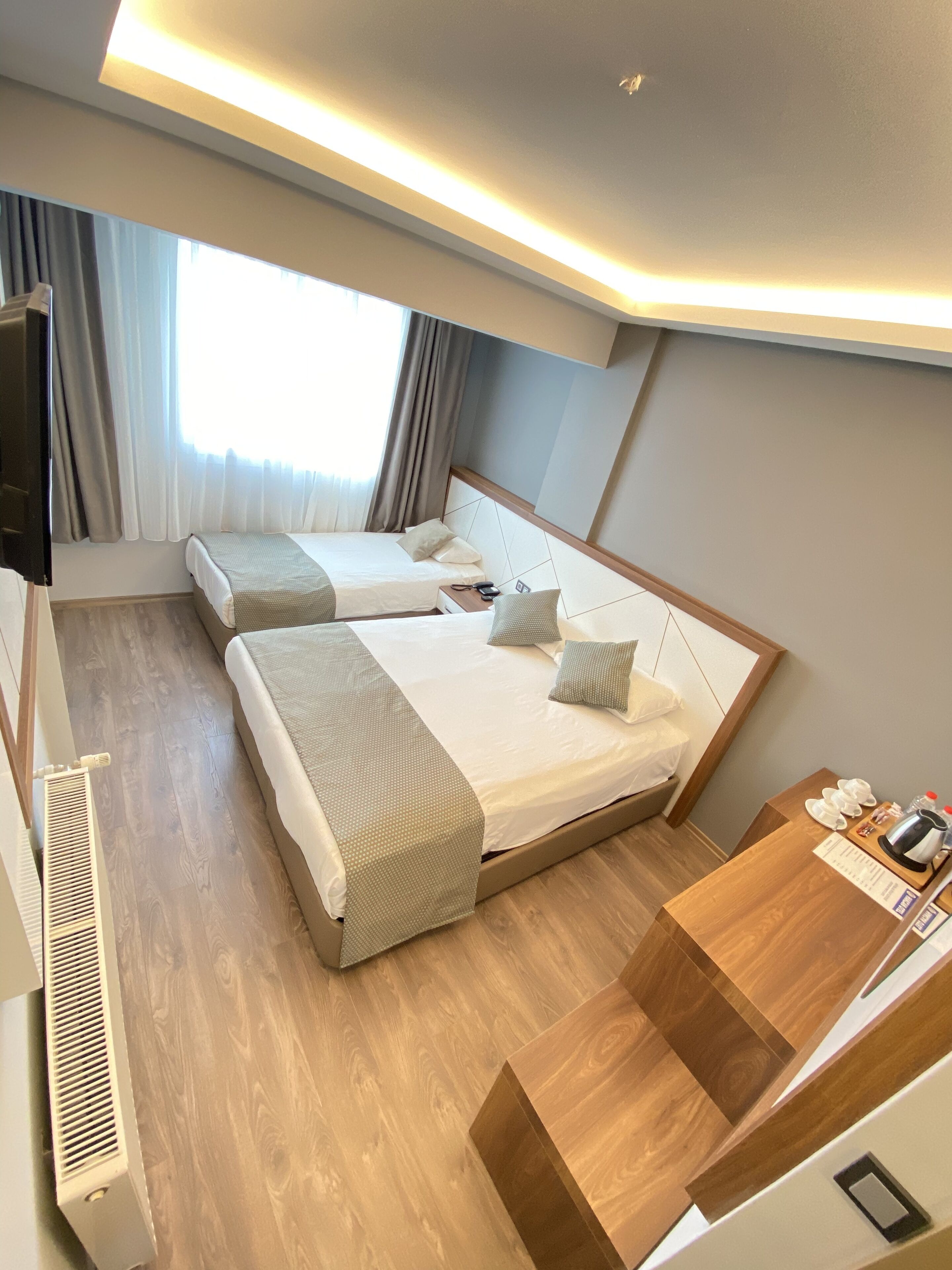 A hotel room with two beds, a wooden floor, a desk with a kettle, and a TV.