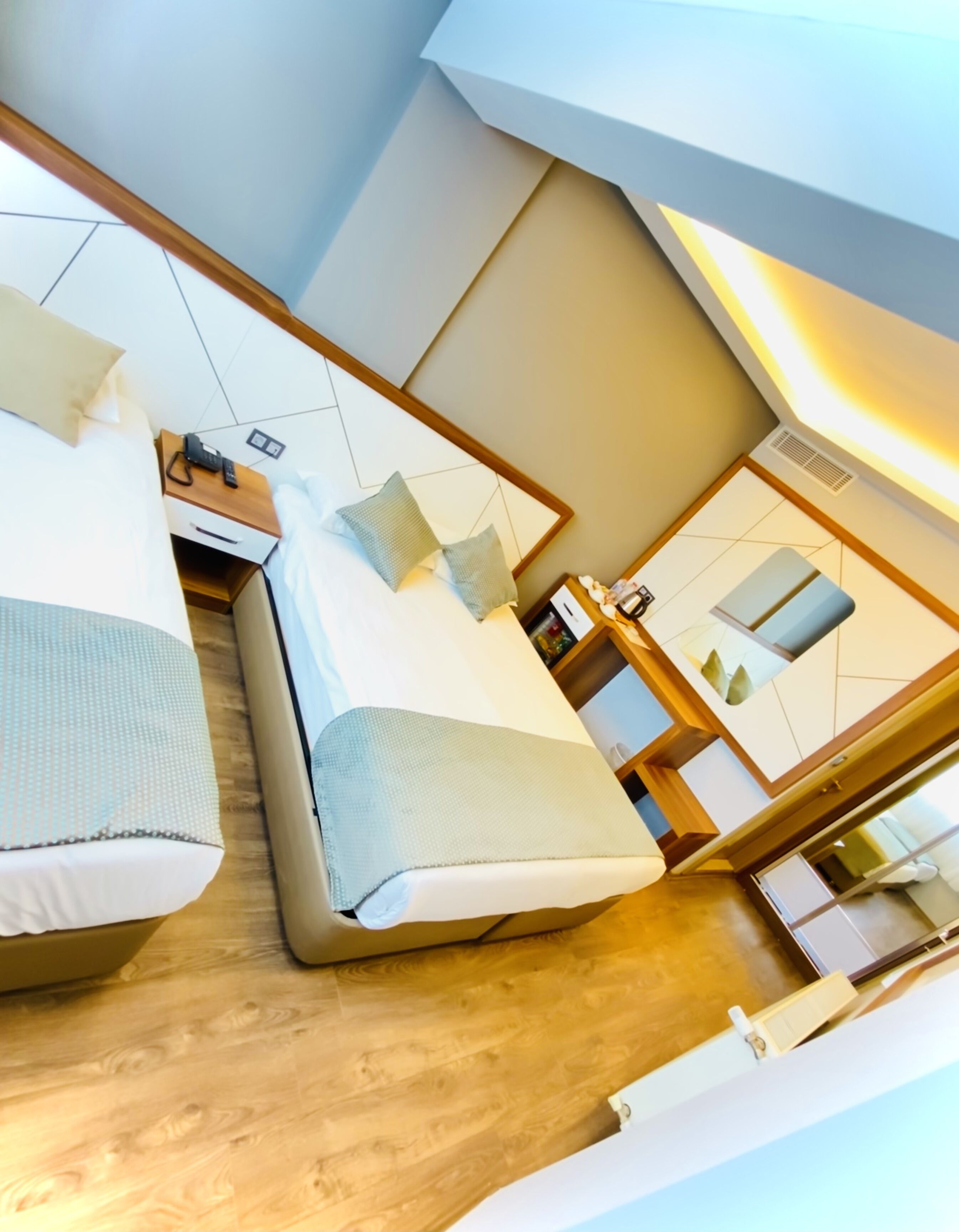 A modern hotel room with two beds, a wooden floor, and a large mirror reflecting the room.