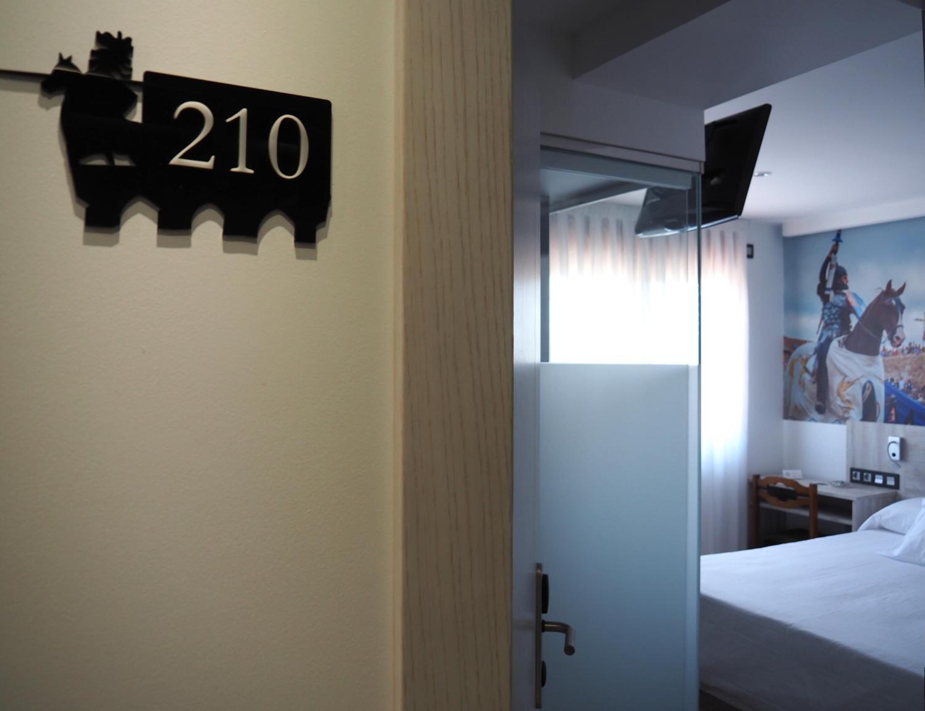 Habitació d'hotel amb llit, una vetust i una televisió penjada a la paret, i una finestra amb cortines.