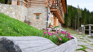 A log cabin with stone walls, a wooden deck, and a flower bed with pink flowers. - Chalet nel Doch (Primiero San Martino di Castrozza)