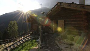A wooden cabin with a stone staircase leading up to a wooden deck, surrounded by mountains and a clear sky. - Chalet nel Doch (Primiero San Martino di Castrozza)