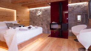 A cozy bedroom with a wooden ceiling, a stone wall, a fireplace, a bed with white linens, and two white chairs. - Chalet nel Doch (Primiero San Martino di Castrozza)