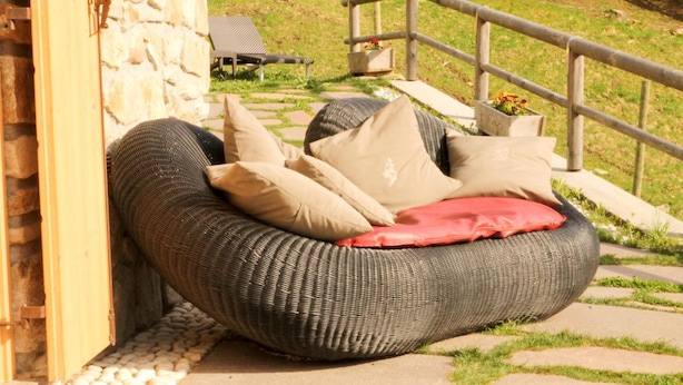 A wicker bean bag chair with cushions, positioned near a stone wall and a wooden railing, overlooking a grassy area. - Chalet nel Doch (Primiero San Martino di Castrozza)