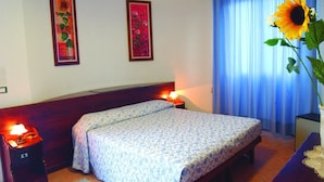 Een hotelkamer met een bed, twee nachtkastjes met lampen, een groot raam met gordijnen, en twee bloemschilderijen in lijsten aan de muur.