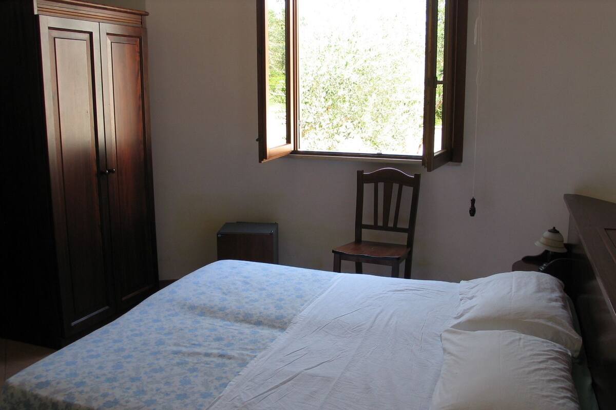Een slaapkamer met een bed, een stoel, een nachtkastje, een raam en een kledingkast.