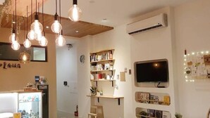 A modern living room with a wooden dining table, clear plastic chairs, a bookshelf, a flat-screen TV, and a ceiling-mounted air conditioning unit. - Warm & Cozy Inn (Hengchun)