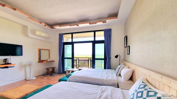 A hotel room with two beds, a TV, a desk, and a balcony with a view. - Warm & Cozy Inn (Hengchun)