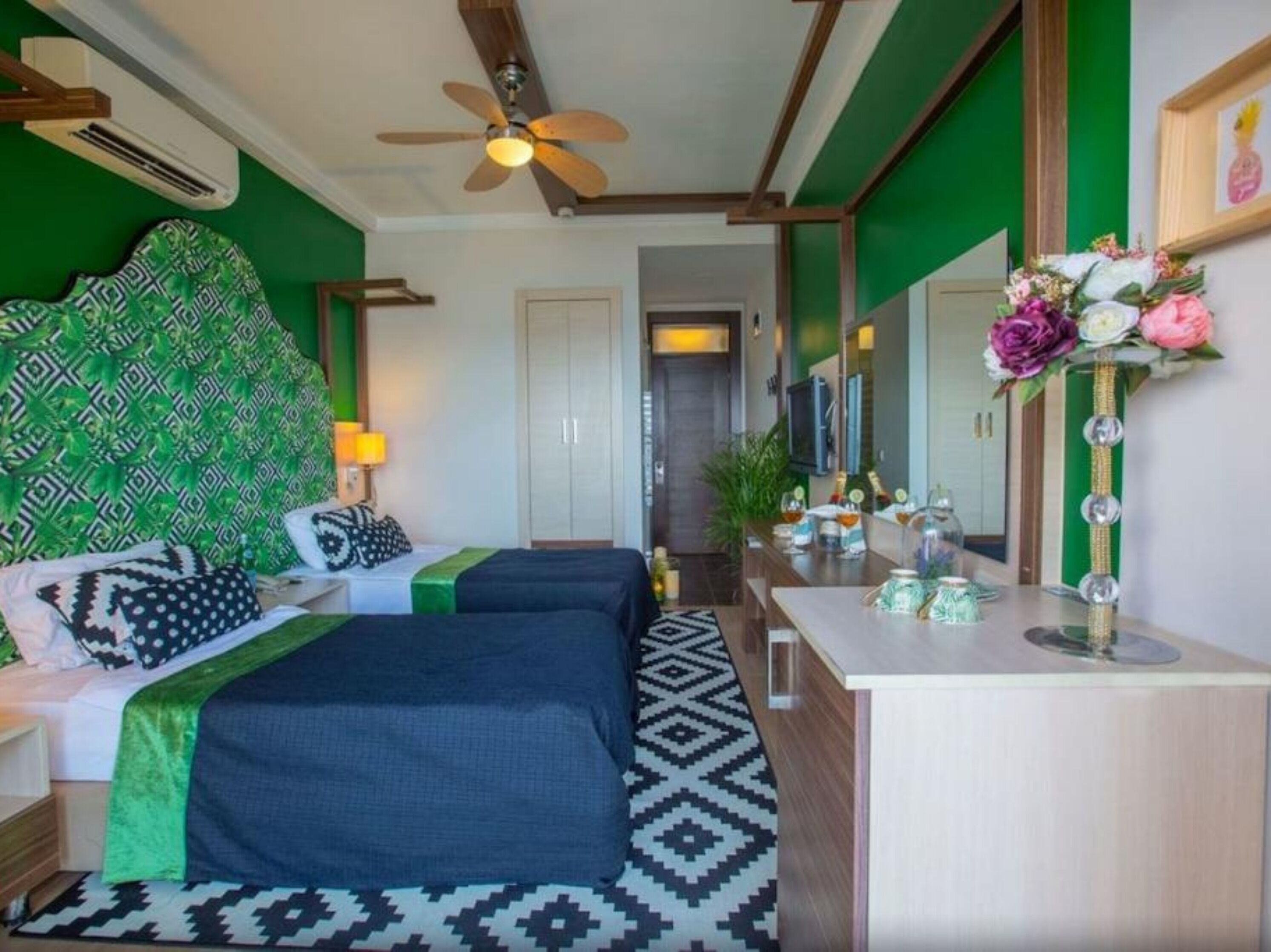 A hotel room with two beds, a ceiling fan, a bedside table with a lamp, a mirror, and a small table with decorative items.