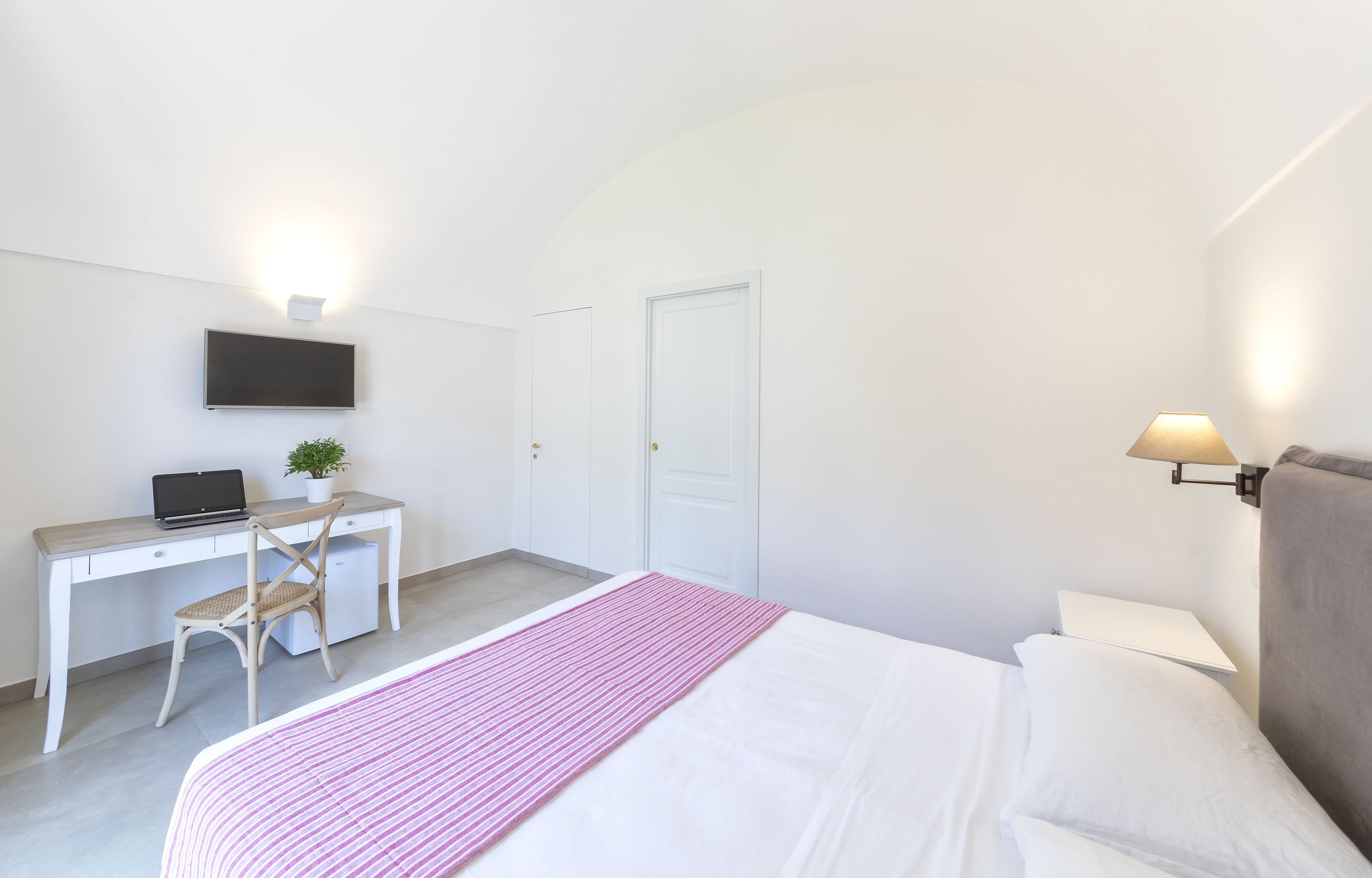 Un dormitorio con cama, escritorio con computadora portátil, silla, planta pequeña y televisor montado en la pared.