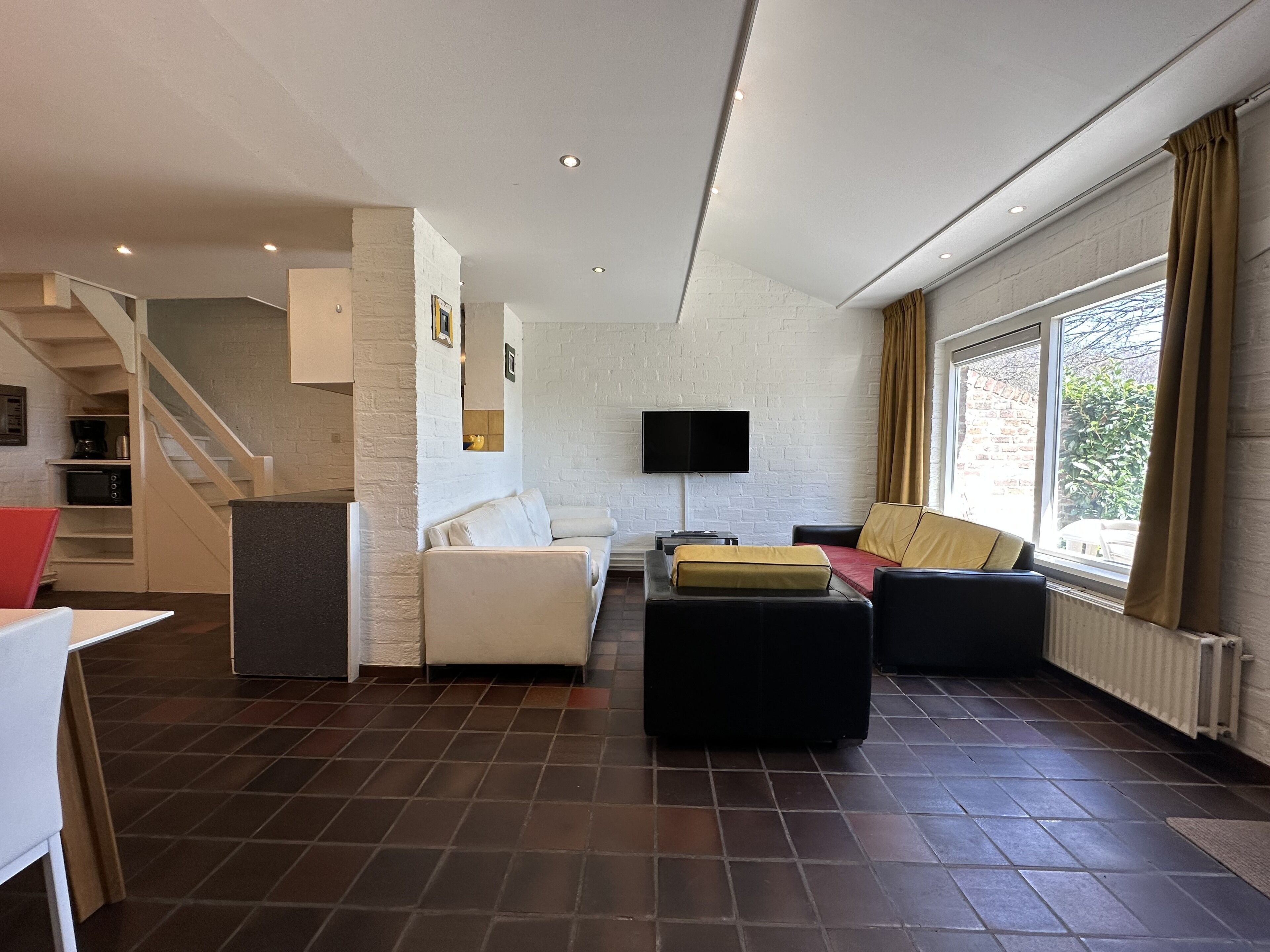 Ein modernes Wohnzimmer mit einer Couch, einem Hocker und einem Flachbildfernseher, der an einer weißen Ziegelwand montiert ist. Im Hintergrund ist eine Treppe zu einer oberen Ebene zu sehen.