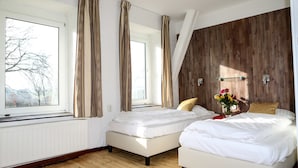 Ein Schlafzimmer mit zwei Betten, großen Fenstern, Holzvertäfelung an einer Wand und einer Blumenvase auf dem Nachttisch.