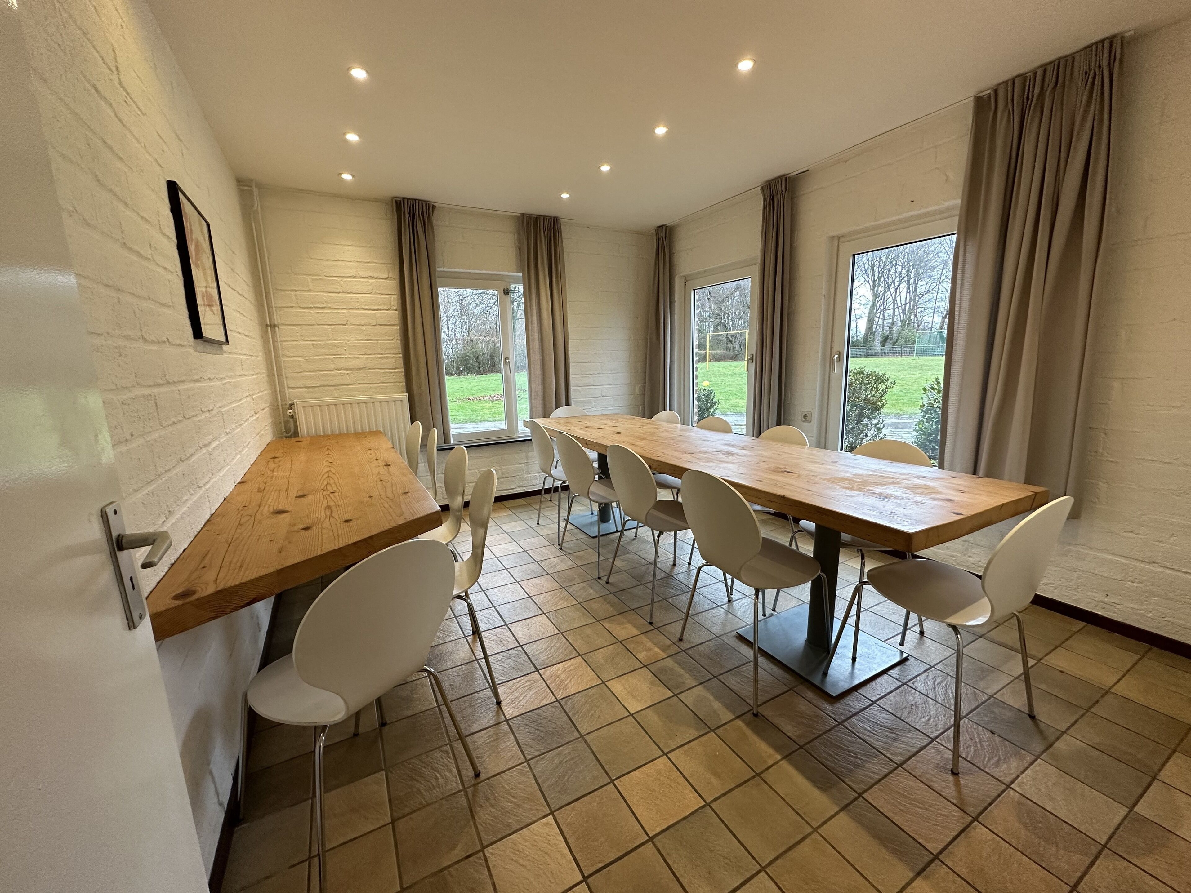 Een eetgedeelte met een lange houten tafel en witte stoelen, een witte bakstenen muur en grote ramen met gordijnen.