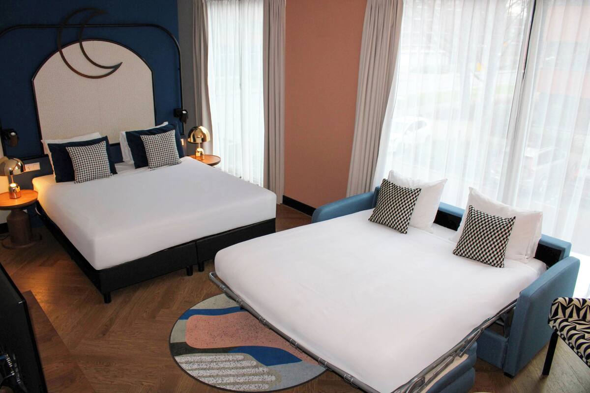Habitación de hotel con dos camas, un ventanal con cortinas translúcidas, un sillón azul y una alfombra redonda.