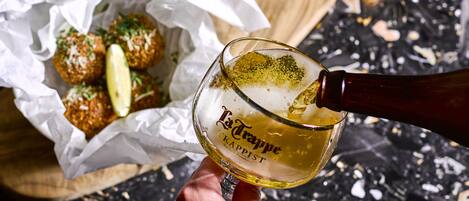 A hand pouring a drink into a glass labeled "La Trappe Prist." In the background, there is a basket with food items and a small bowl of sauce.