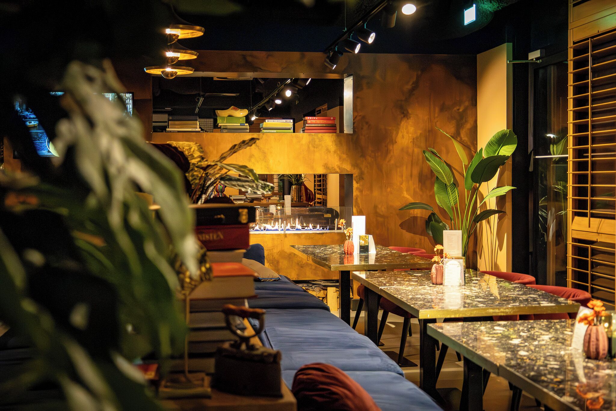 En hyggelig restaurant med et langt fællesskabslaug, en reol med bøger og mange planter.