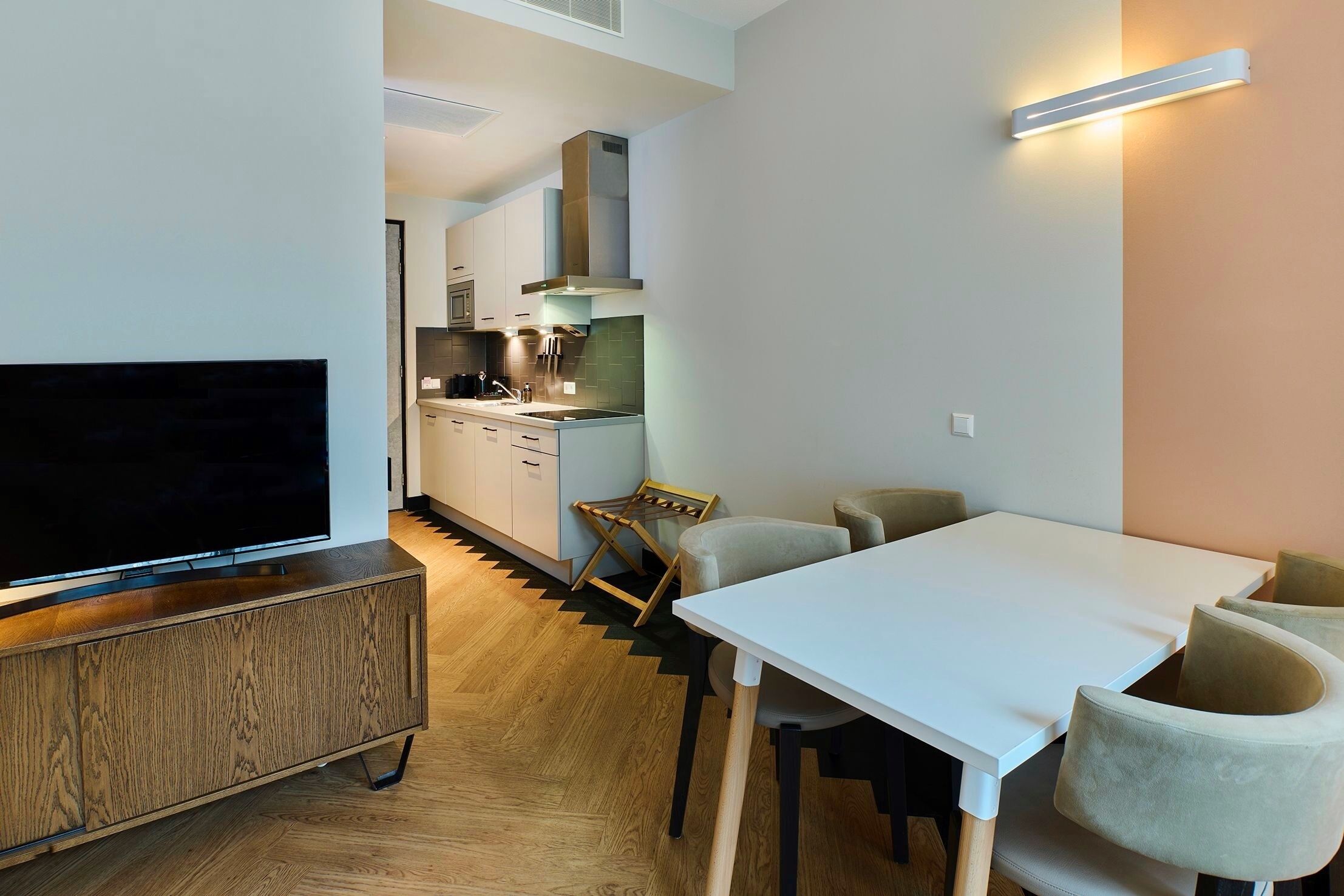 Una cocina moderna con estufa integrada, un comedor con mesa y sillas blancas, y un televisor sobre un mueble de madera.