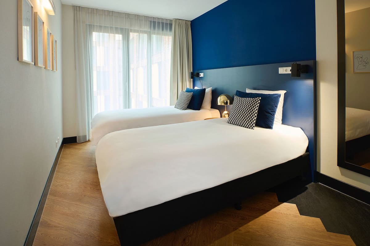 Habitación de hotel con dos camas, un ventanal con cortinas translúcidas, una pared de acento azul y una lámpara moderna.