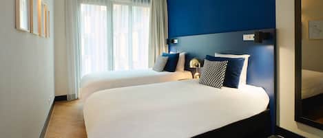 A hotel room with two beds, a large window with sheer curtains, a blue accent wall, and a modern lamp.