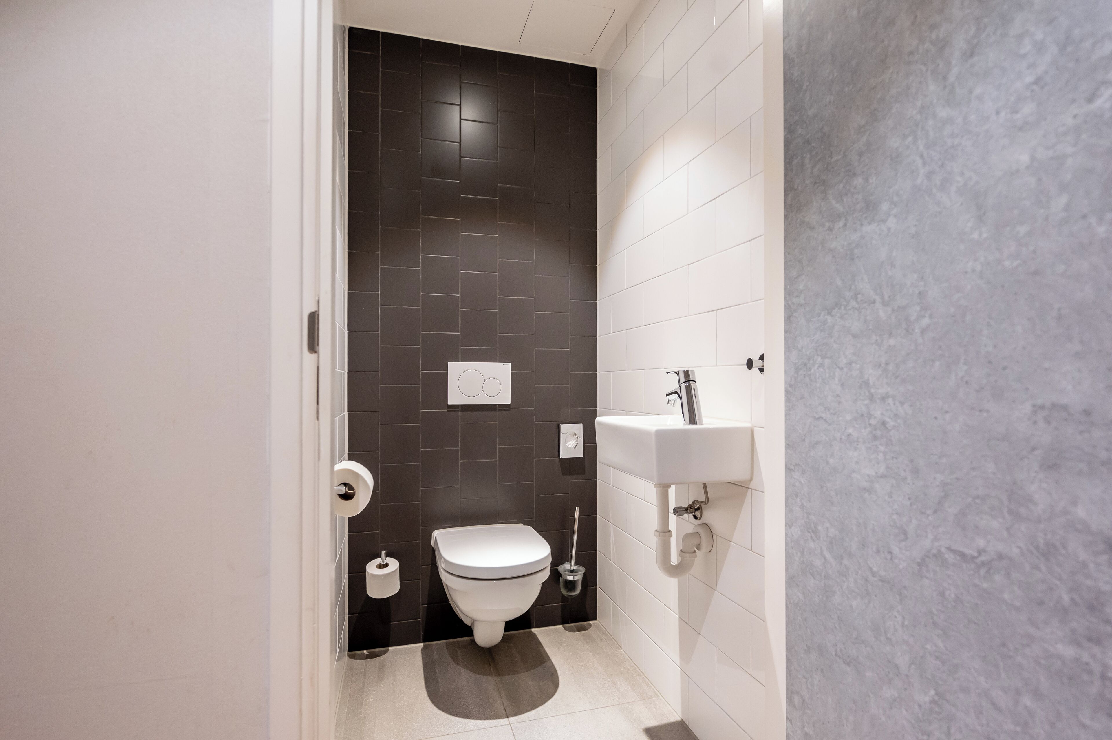 Un baño moderno con pared de azulejos oscuros, un inodoro blanco, un lavamanos y un dispensador de papel higiénico.