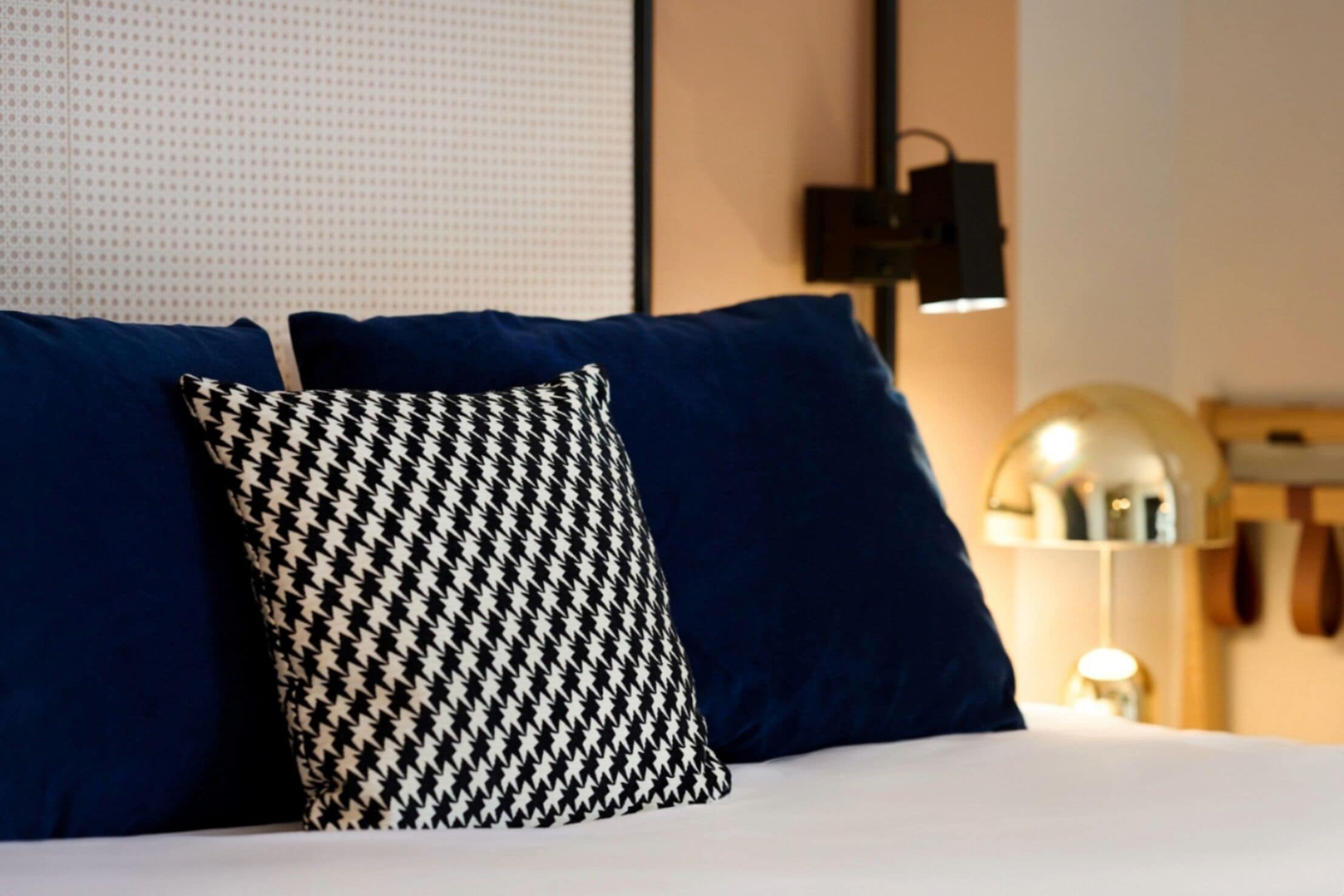 A bed with blue and houndstooth patterned pillows, a wall-mounted light, and a partial view of a wooden shelf.