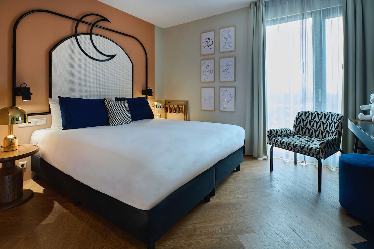 Una habitación de hotel moderna con una cama grande, piso de madera, un sillón estampado y obras de arte en las paredes.