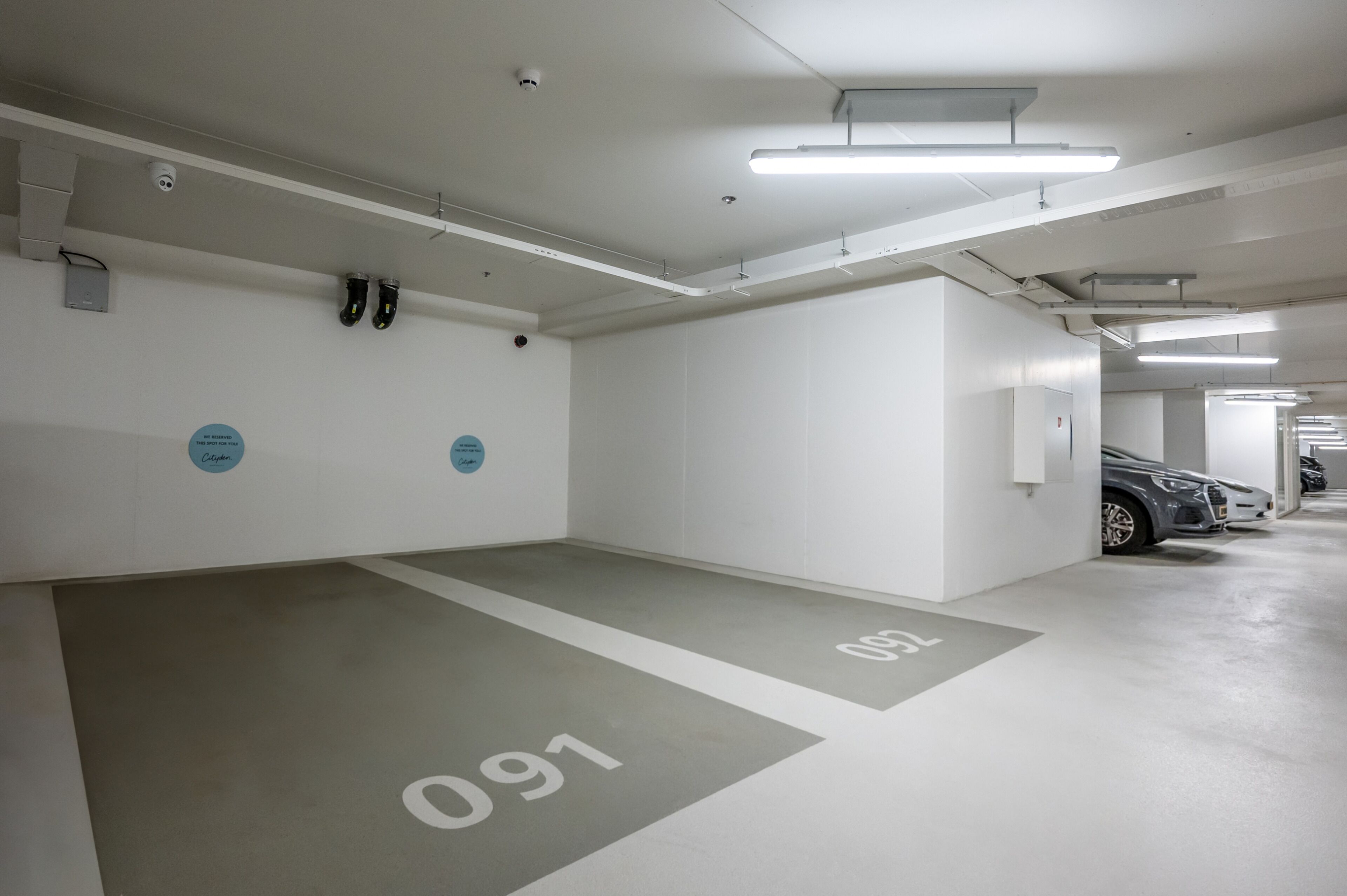 Un parking propre et moderne, avec des murs et des sols blancs, des places de stationnement clairement délimitées et un éclairage fluorescent au plafond.