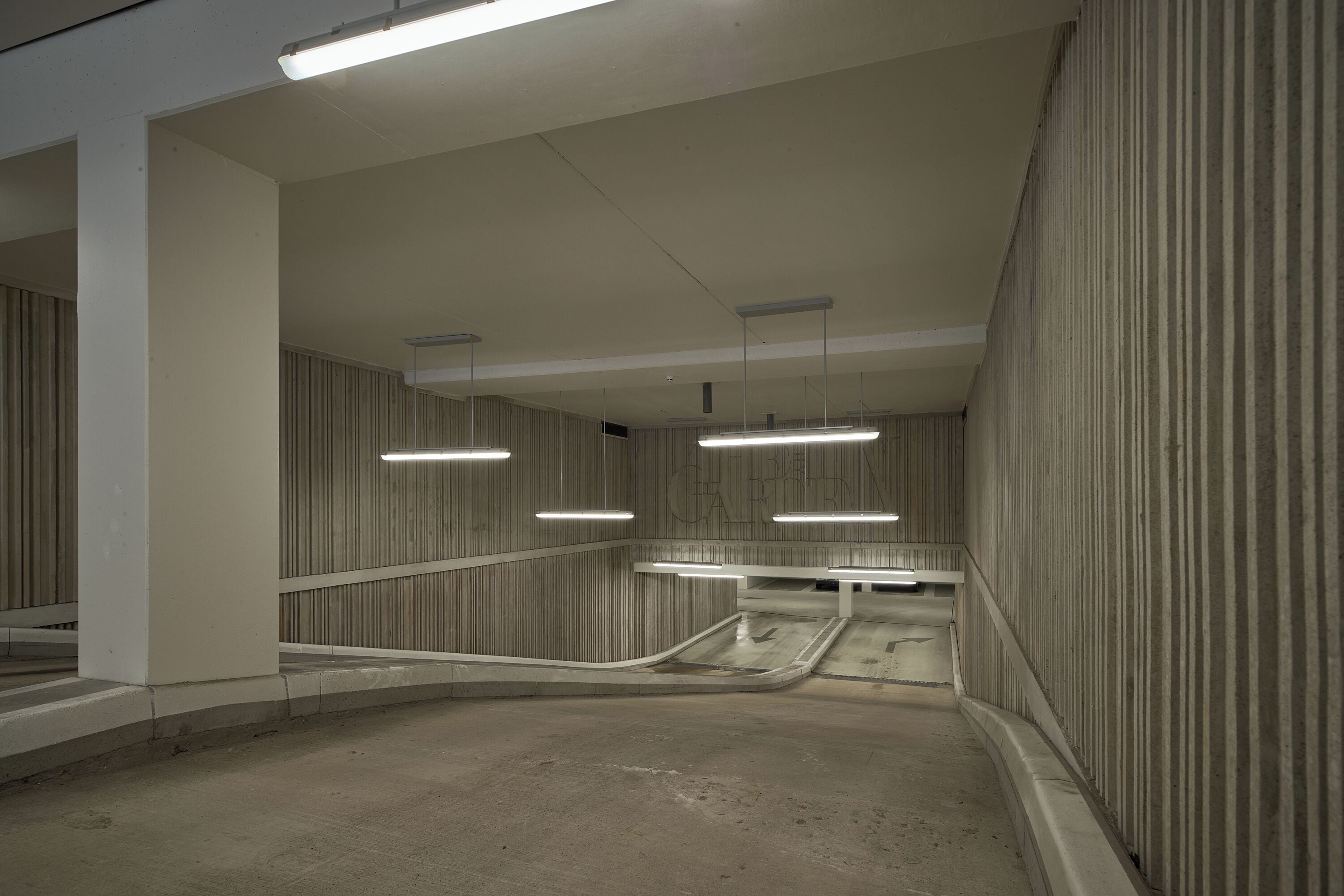 Eine moderne, leere Tiefgarage mit Betonböden und -wänden, die eine zentrale Rampe und Deckenbeleuchtung mit Leuchtstoffröhren aufweist.