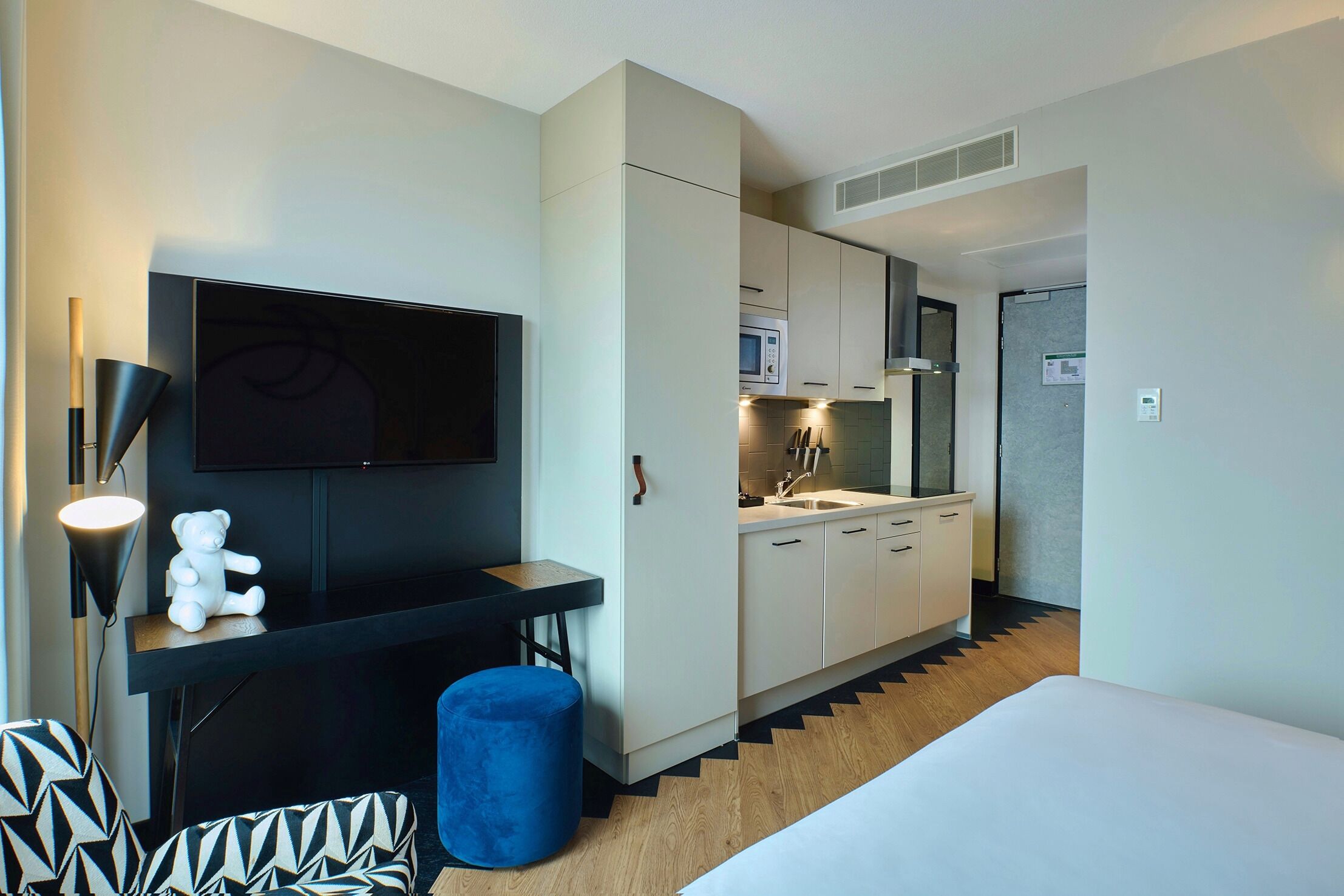 A compact hotel room with a kitchenette, a flat-screen TV, a small seating area, and a bed.