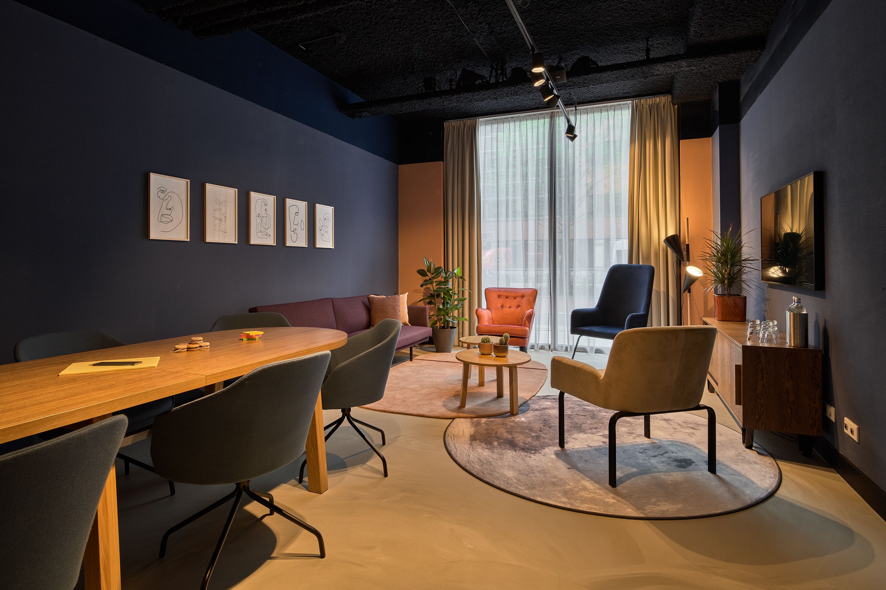 Uma sala de reuniões moderna com mesa de madeira, quatro cadeiras, um sofá e uma janela grande com cortinas.