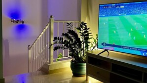 Een woonkamer met een televisie waarop een voetbalwedstrijd wordt vertoond, een trap en een potplant.