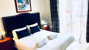 Een keurig opgemaakt bed met witte lakens en twee opgerolde handdoeken, een nachtkastje met een lamp, een ingelijst foto aan de muur, en een groot raam met gordijnen.