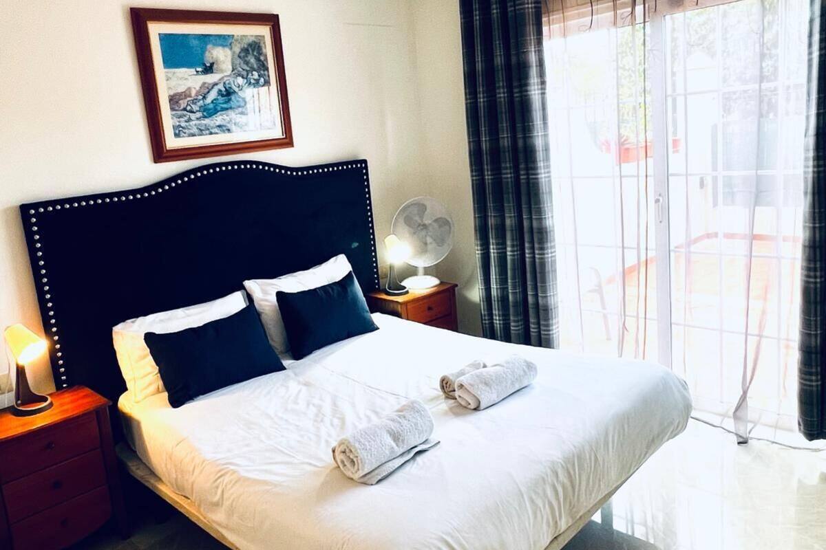 Een keurig opgemaakt bed met witte lakens en twee opgerolde handdoeken, een nachtkastje met een lamp, een ingelijst foto aan de muur, en een groot raam met gordijnen.