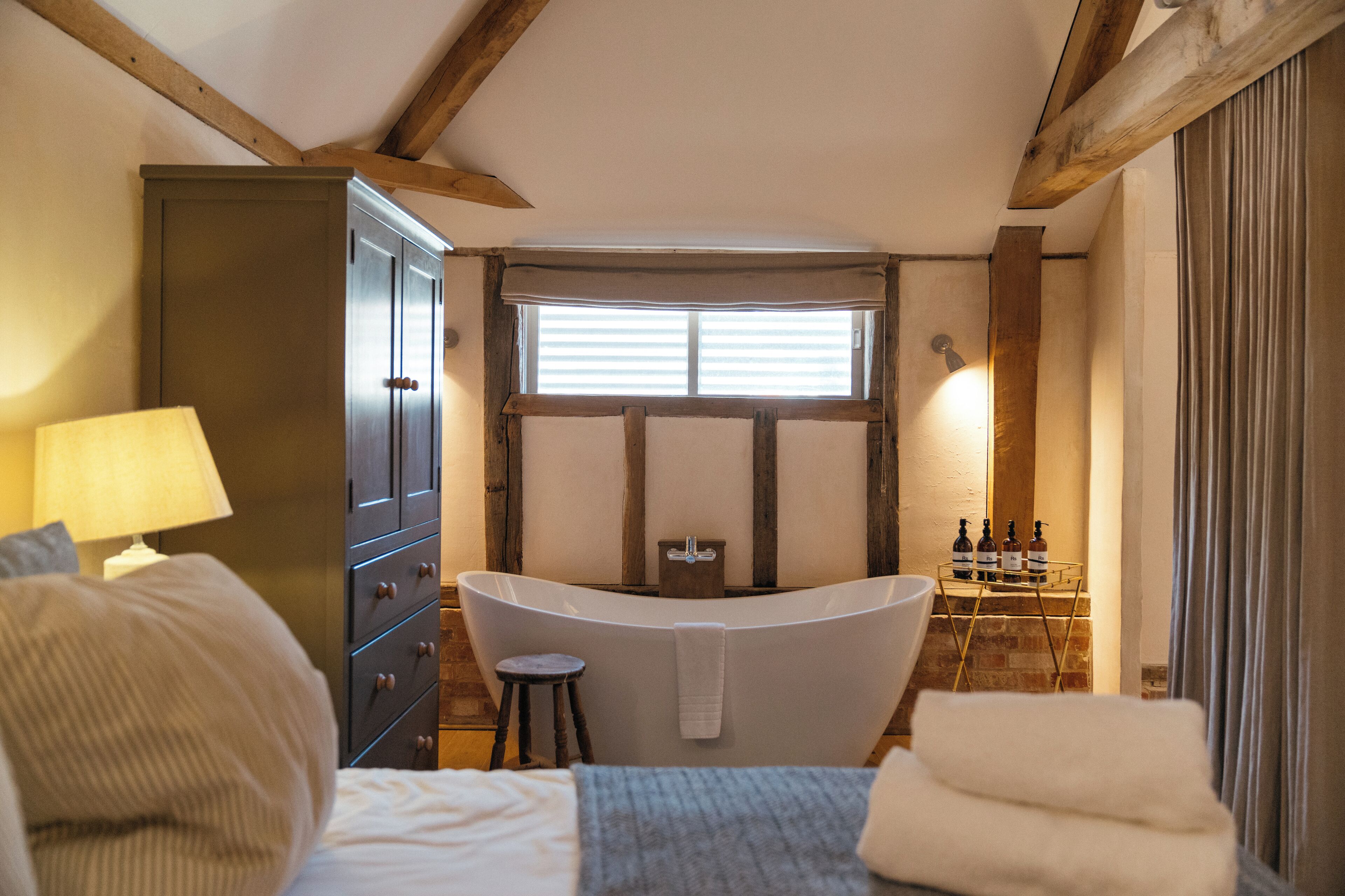 A bedroom with a large bed, a wooden wardrobe, a bathtub, and a window with blinds.