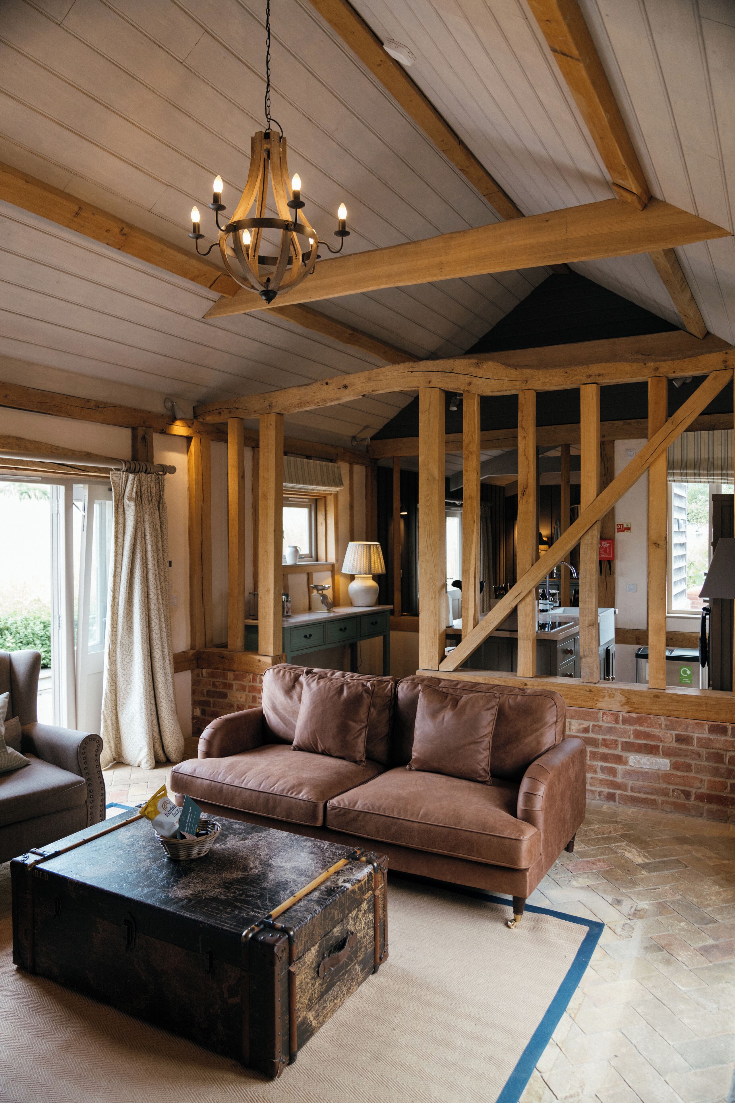 A rustic interior with exposed wooden beams, a leather sofa, a vintage trunk as a coffee table, and a chandelier.