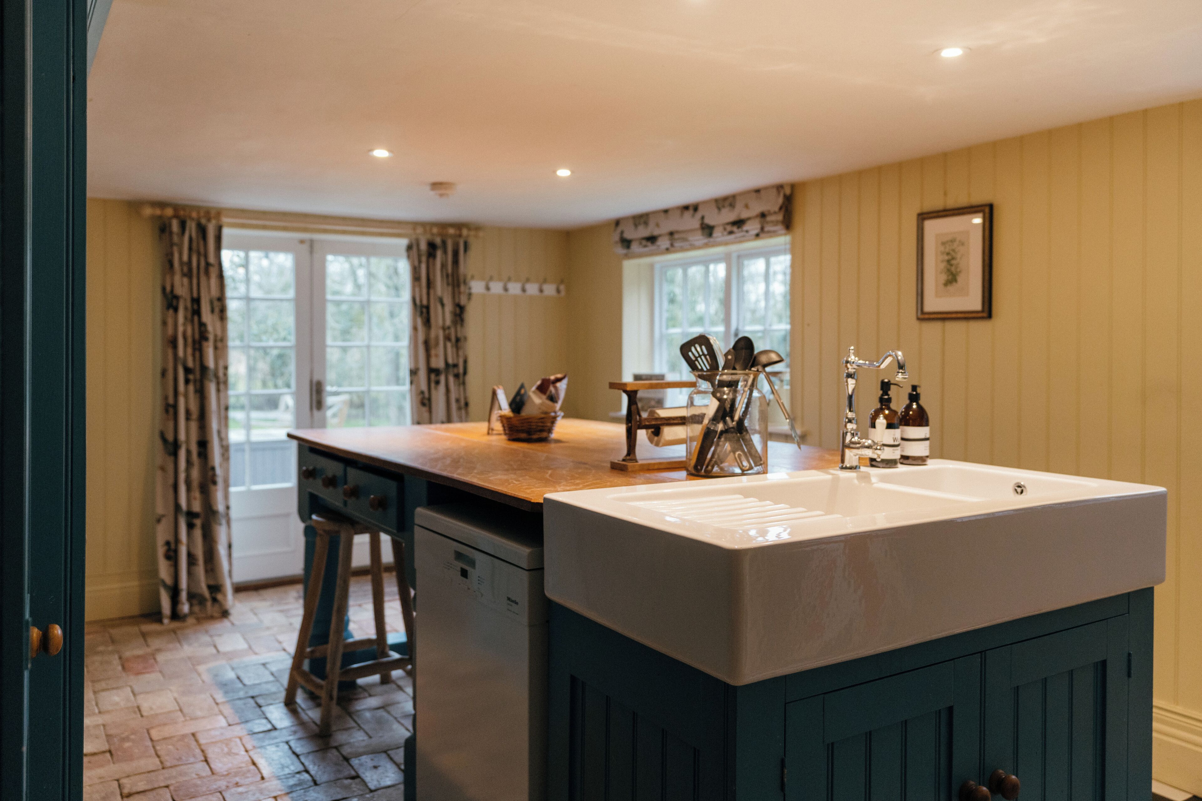 A kitchen with a large island, wooden stools, a sink, and a window with curtains.