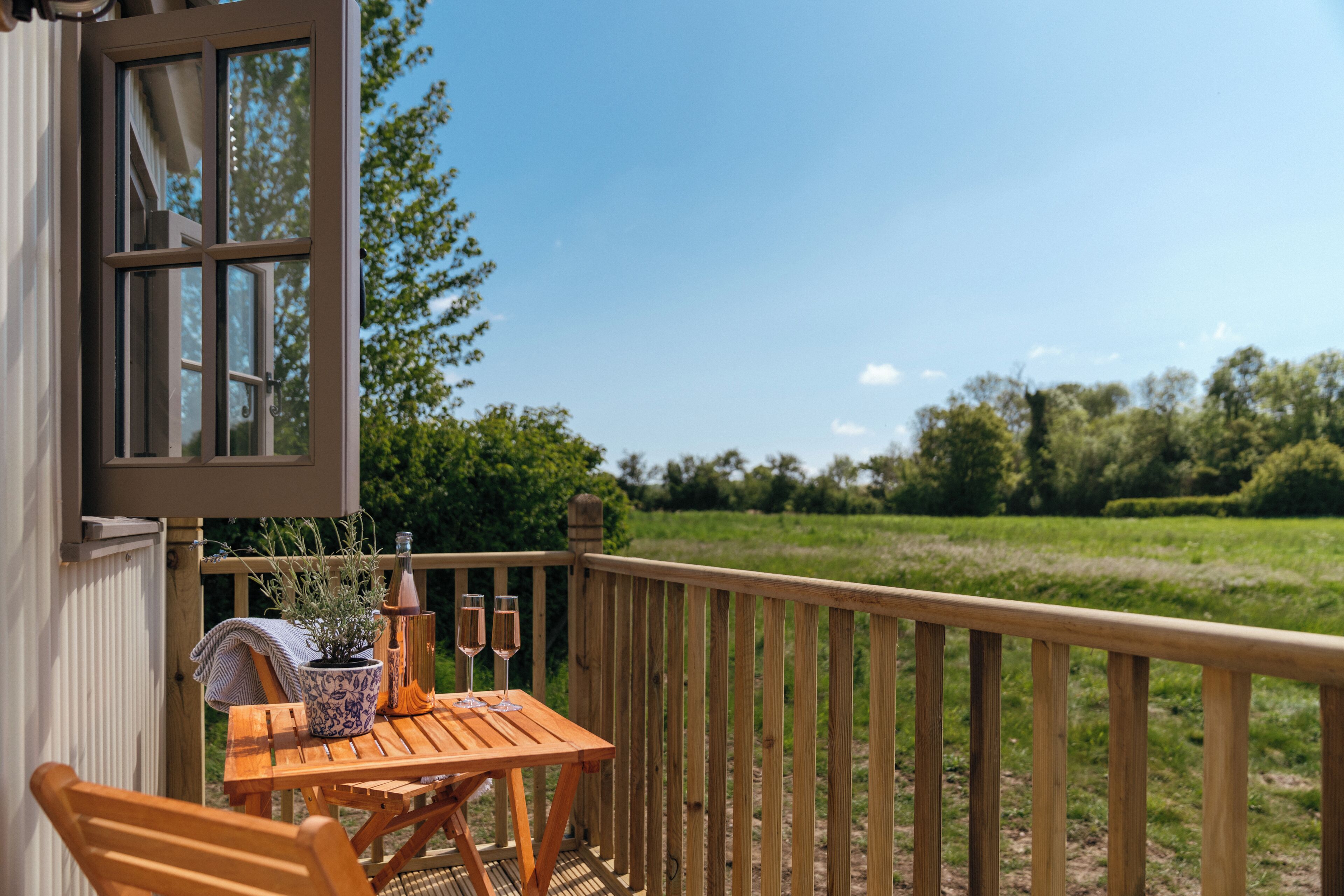 A wooden deck with a table and chairs, a potted plant, and two glasses of champagne.