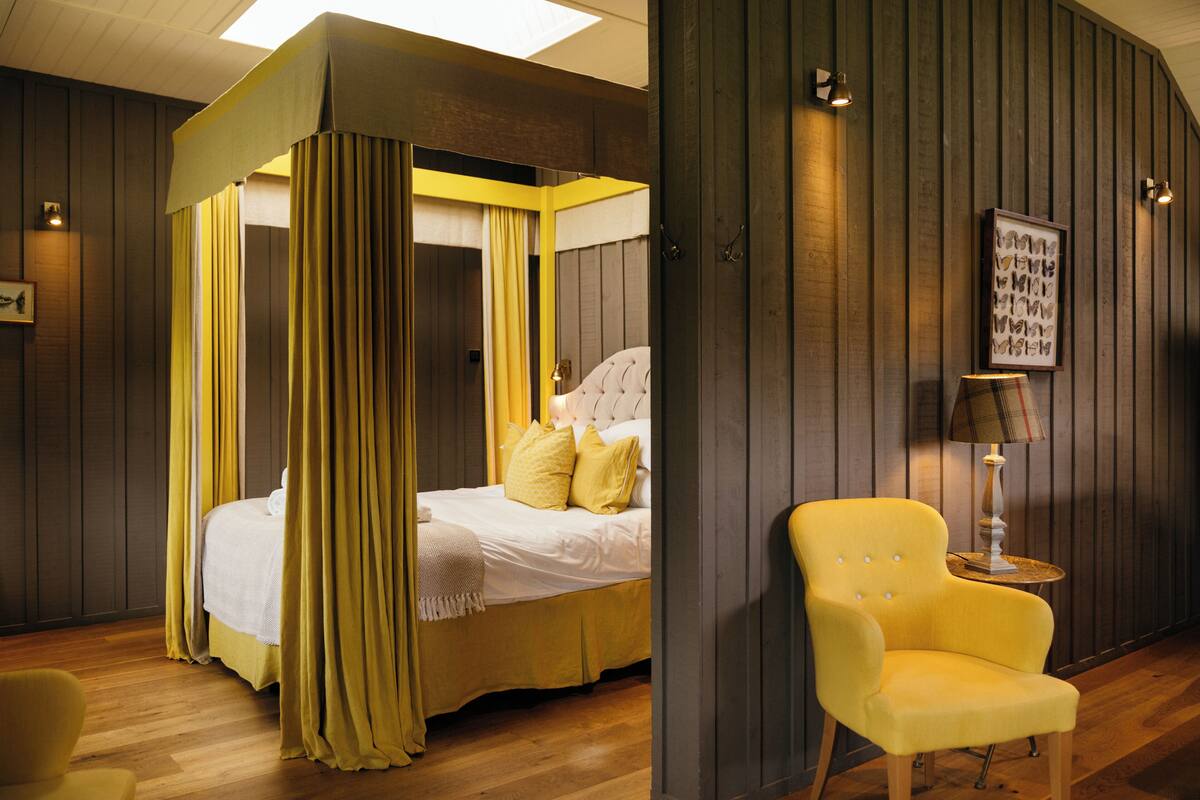 A bedroom with a four-poster bed, a yellow armchair, and a bedside table with a lamp.
