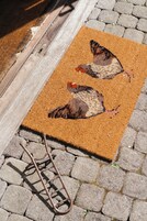 A doormat with a bird design, placed on a cobblestone surface next to a wooden door.